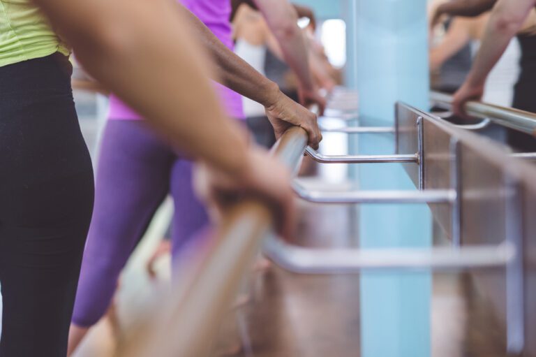 A multiethnic group of young women do a barre workout together at a modern gym. They are gripping the barre with one hand. The shot is from behind them and is focused on their hands as they grip the barre.